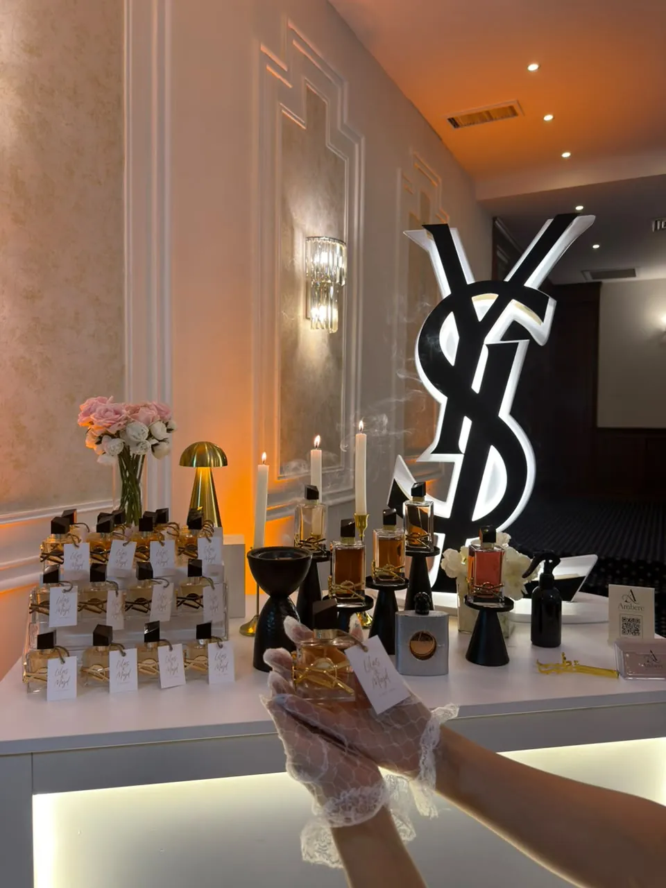 يد بقفاز دانتيل بتقدّم عطر قدام بوث Ambere، وعلى البوث شموع وعطور ومبخرة