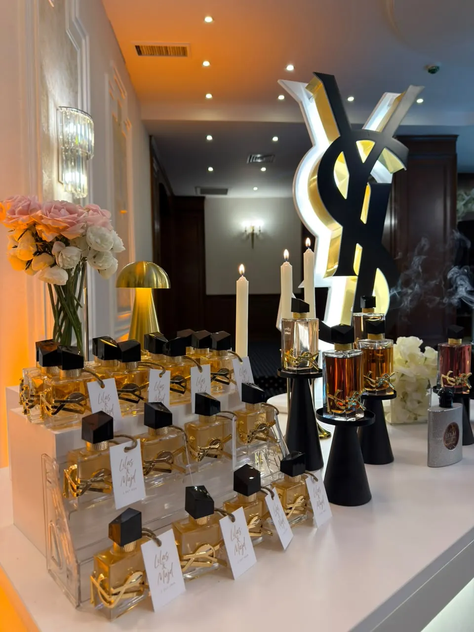 مدرّج توزيعات عطور بكروت أسماء العروسين، وخلفه شموع وعطور على البوث